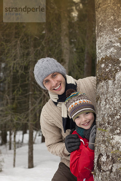 Porträt eines glücklichen Vaters und Sohnes hinter Baumstamm in verschneiten Wäldern