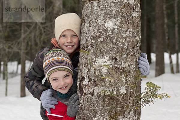 Portrait von glücklichen Jungen hinter Baumstamm in verschneiten Wäldern