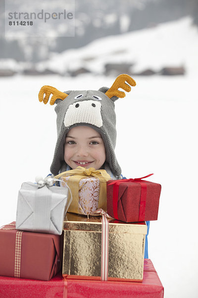 Porträt eines lächelnden Jungen mit Rentierhut und einem Stapel Weihnachtsgeschenke im Schnee.