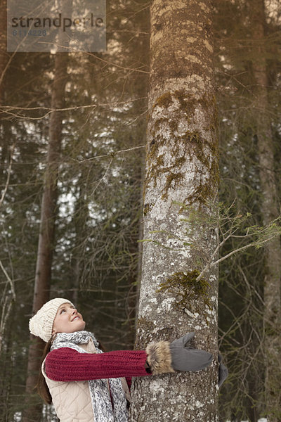 Lächelnde Frau umarmt Baumstamm im Wald