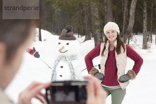 Mann fotografiert lächelnde Frau mit Händen auf den Hüften neben Schneemann im Wald