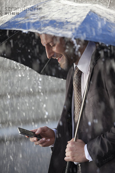 Geschäftsmann SMS auf dem Handy unter dem Schirm bei Regen