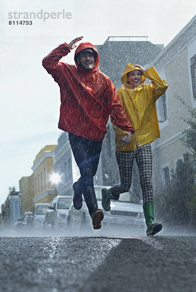 Glückliches Paar in Regenmänteln  die im Regen die Straße hinunterlaufen