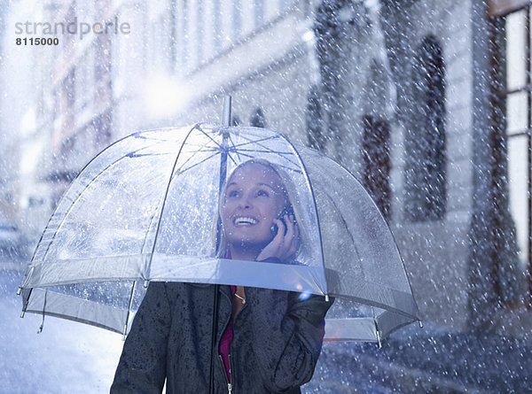 Lächelnde Geschäftsfrau beim Telefonieren unter dem Regenschirm in der verregneten Straße