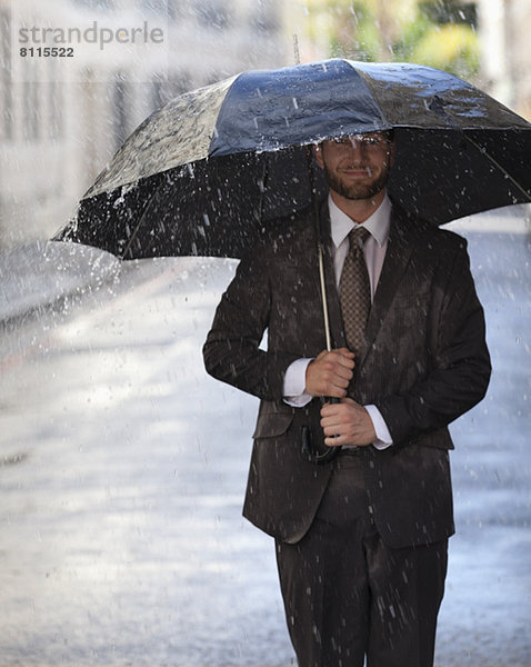 Glücklicher Geschäftsmann mit Regenschirm  der in einer verregneten Straße spazieren geht.