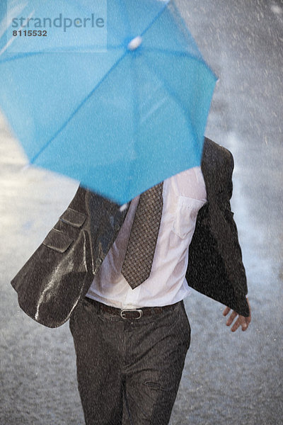 Geschäftsmann mit winzigem Regenschirm  der in einer verregneten Straße läuft.