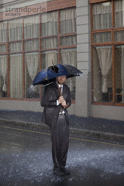 Geschäftsmann steht unter zerbrochenem Regenschirm in einer verregneten Straße.