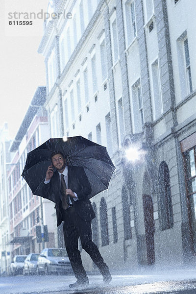 Geschäftsmann im Gespräch auf dem Handy unter dem Regenschirm in der verregneten Straße