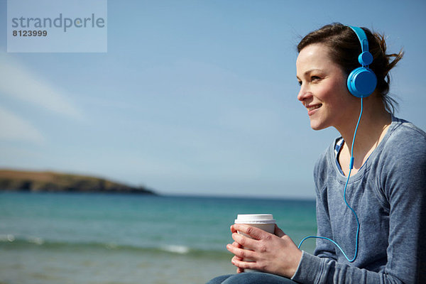 Junge Frau an der Küste mit Kaffee und Kopfhörer