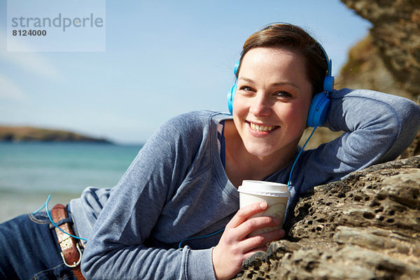 Porträt einer jungen Frau an der Küste mit Kaffee und Kopfhörer