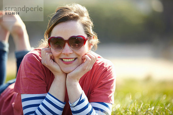 Porträt einer jungen Frau mit herzförmiger Sonnenbrille auf Gras liegend