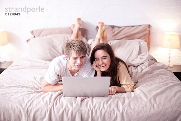 Junges Paar liegt auf dem Bett und schaut auf den Laptop.