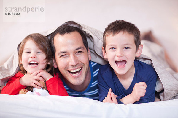 Vater und zwei kleine Kinder posieren unter der Bettdecke