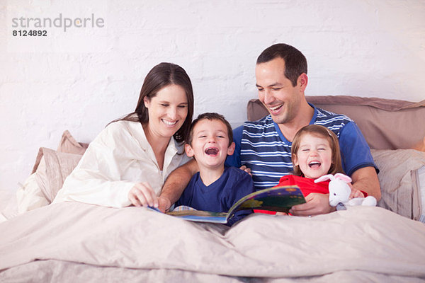 Eltern und zwei kleine Kinder schauen sich das Bilderbuch im Bett an.