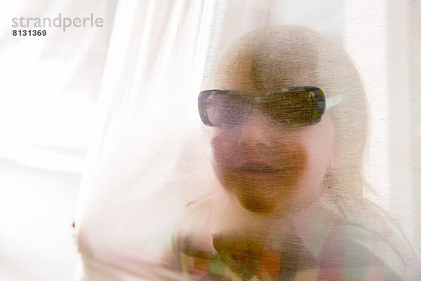 Mädchen mit Sonnenbrille durch den Vorhang schauend