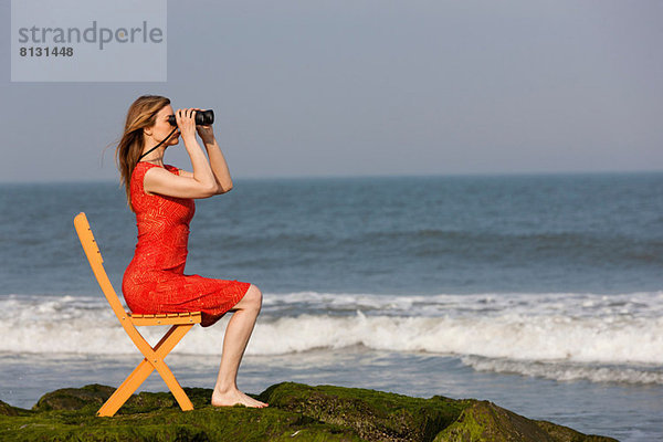 Reife Frau sitzt auf einem Stuhl am Strand mit Fernglas