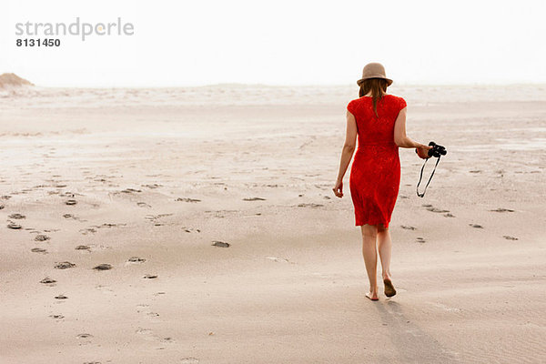 Reife Frau in rotem Kleid  die am Strand spazieren geht.