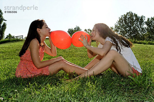 Zwei junge Frauen sitzen auf Gras und verbeugen sich mit roten Luftballons.
