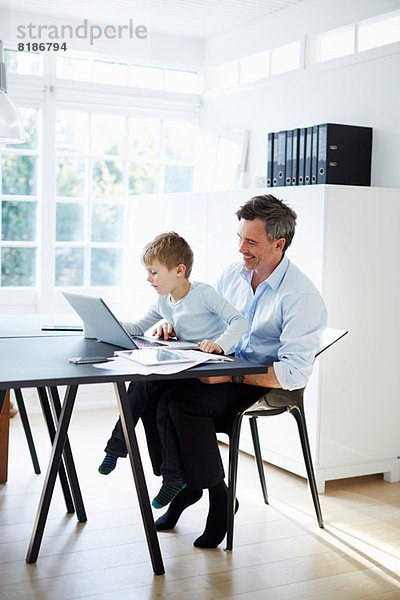 Mann am Schreibtisch sitzend mit Sohn auf dem Knie mit Laptop und Papierkram