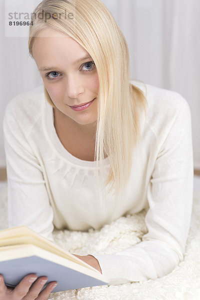 Porträt einer jungen Frau  die auf der Couch liegt und ein Buch liest  lächelnd.