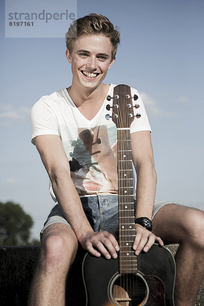 Deutschland  Junger Mann im Park sitzend  Gitarre haltend