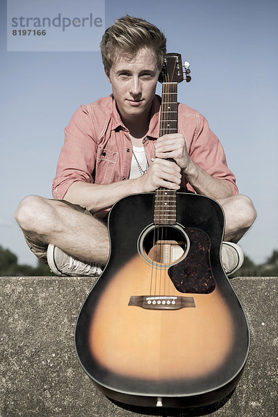 Germany  Young man sitting on wall with guitar