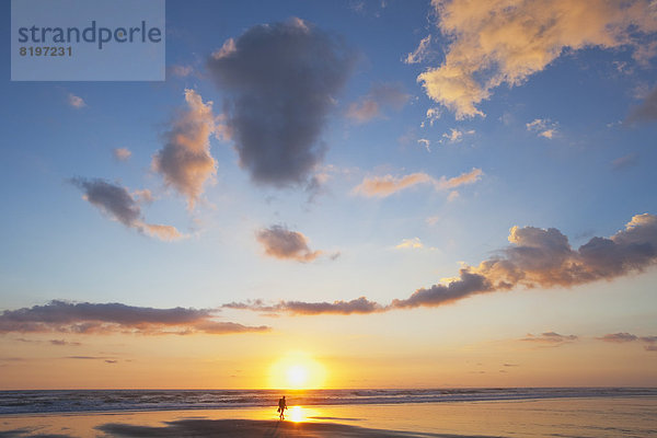 Neuseeland  Blick auf einen Mann mit Rucksack  der bei Sonnenuntergang am Strand entlang läuft.