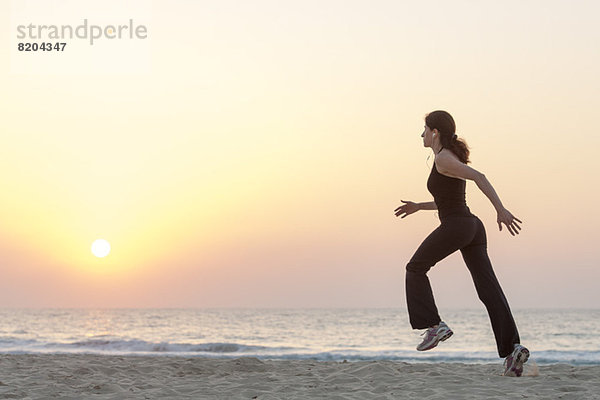 Frau beim Joggen am Strand bei Sonnenaufgang mit Kopfhörer  Seitenansicht