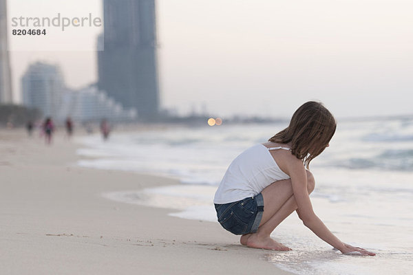 Mädchen kauernd am Strand mit Wasser spielen  Seitenansicht