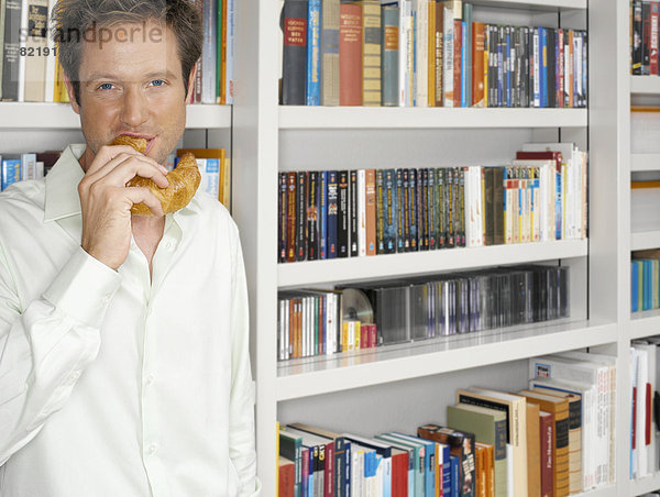 Mann steht vor einem Bücherregal und isst ein Croissant