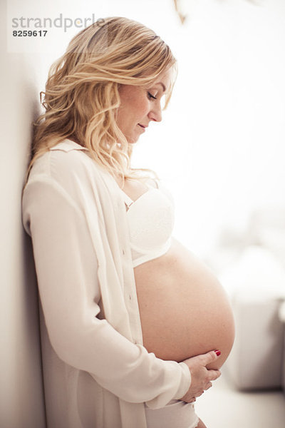Seitenansicht einer schwangeren Frau  die den Bauch berührt  während sie sich zu Hause an die Wand lehnt.