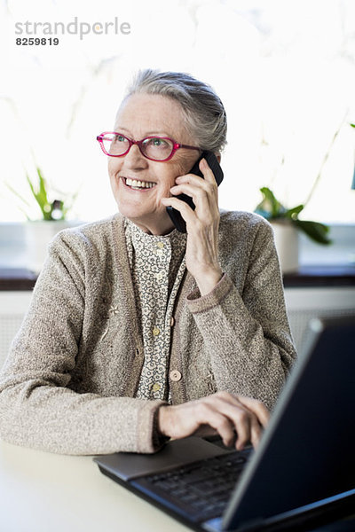 Fröhliche Seniorin mit Handy im Sitzen mit Laptop auf dem Tisch