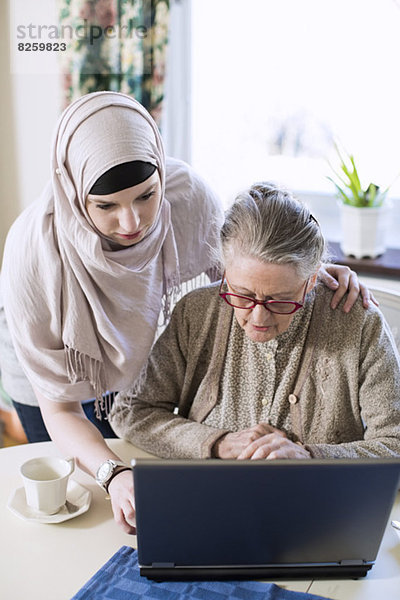 Seniorin mit häuslicher Betreuerin mit Laptop am Tisch