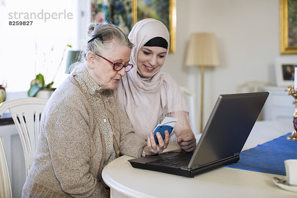 Junge Heimpflegerin mit älterer Frau  die mit dem Laptop zu Hause in die Bank geht.