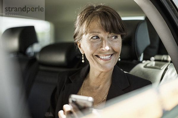 Glückliche Geschäftsfrau  die ein Handy hält und durch das Taxifenster schaut.