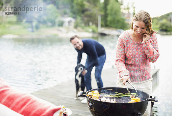 Fröhliche Frau beim Grillen mit Mann und Hund im Hintergrund am Pier