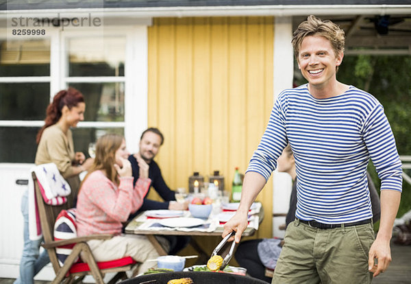 Porträt des glücklichen Mannes beim Grillen mit Freunden am Esstisch im Hintergrund