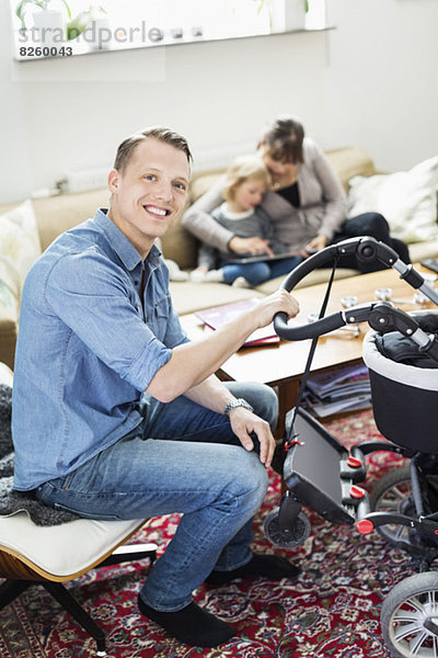 Porträt eines glücklichen Mannes mit Kinderwagen im Wohnzimmer mit Familie im Hintergrund