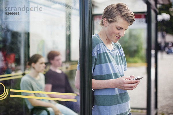 Junger Mann mit Handy beim Warten mit Freunden an der Bushaltestelle