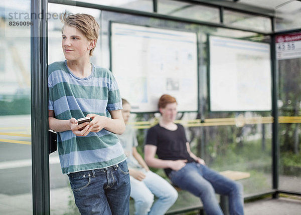 Junger Mann mit Handy beim Warten mit Freunden an der Bushaltestelle