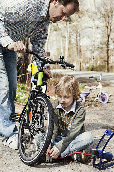 Junge füllt Fahrradreifen mit Fußpumpe  während Vater ihn auf der Straße beobachtet.