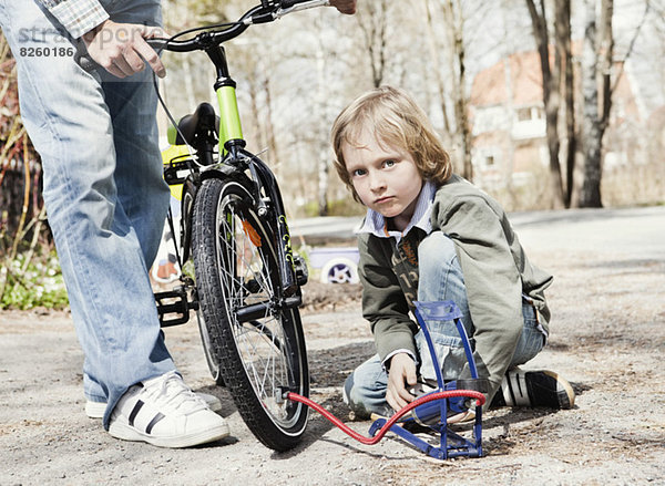 Portrait eines Jungen  der Fahrradreifen mit Fußpumpe füllt  während sein Vater auf der Straße steht.