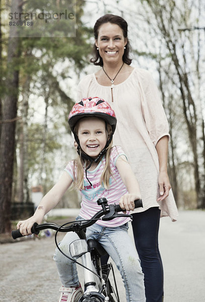 Porträt eines glücklichen Mädchens  das Fahrrad fährt  während die Mutter hinter ihr auf der Straße steht.