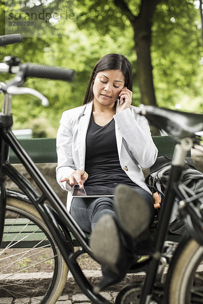 Geschäftsfrau mit digitalem Tablett und Handy auf Parkbank mit Füßen auf dem Fahrrad