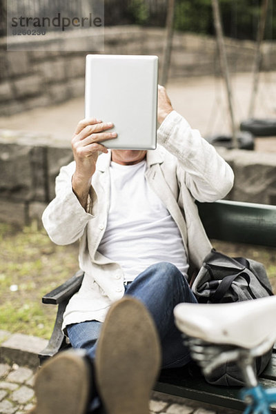 Geschäftsmann hält digitales Tablett vor das Gesicht und entspannt sich auf der Parkbank