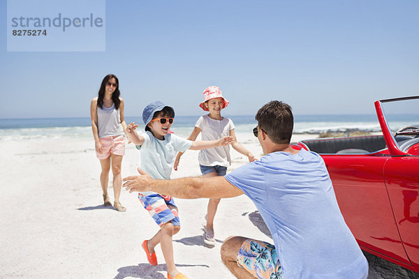 Vater  der die Kinder am Strand neben dem Cabriolet umarmt.