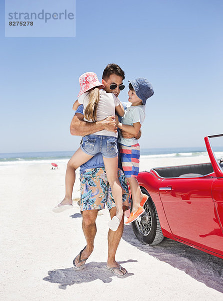 Vater umarmt Kinder am Strand neben dem Cabriolet