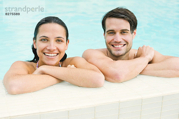 Porträt eines lächelnden Paares  das sich am Rand des Swimmingpools lehnt.
