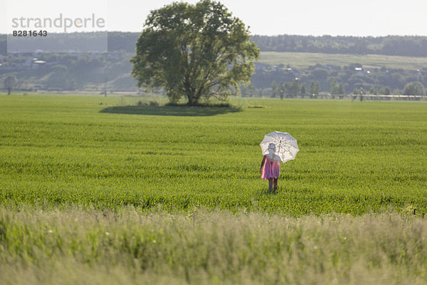 Ein junges Mädchen geht durch ein Feld und benutzt einen Regenschirm als Sonnenschutz.