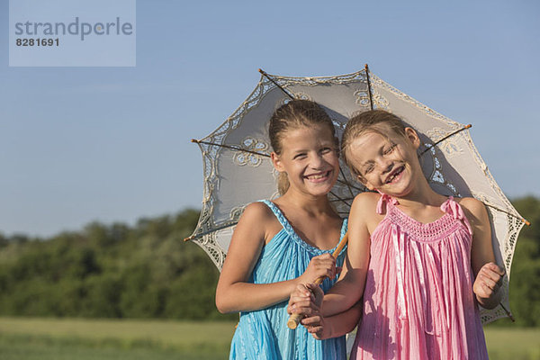 Lachende Zwillingsschwestern stehen auf einem Feld unter einem Schirm.